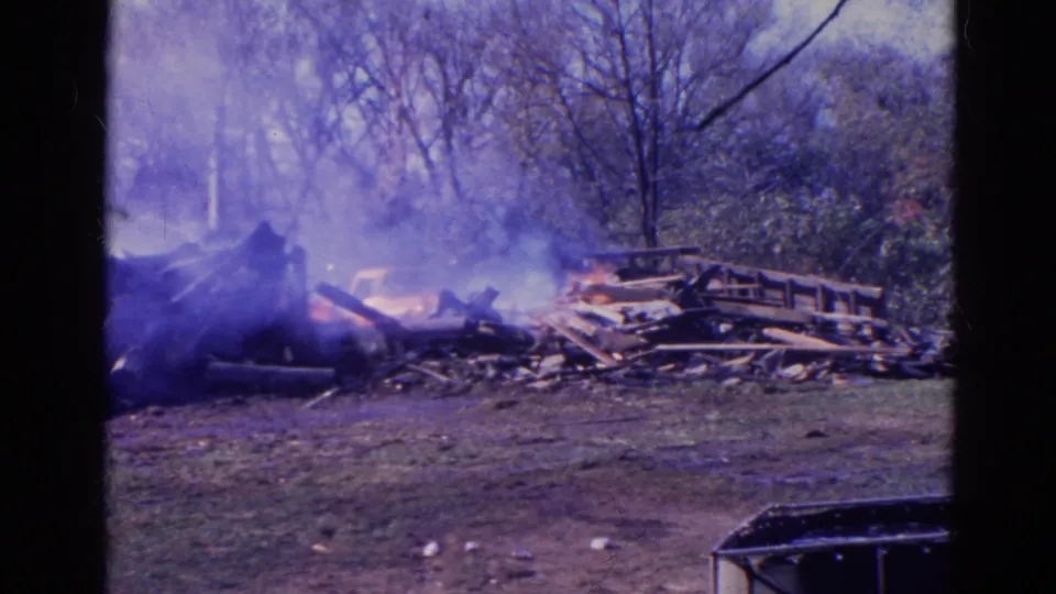 1968 Wauconda, Illinois home-movie frame showing smoke and flames around a debris pile with stacked wood at right.