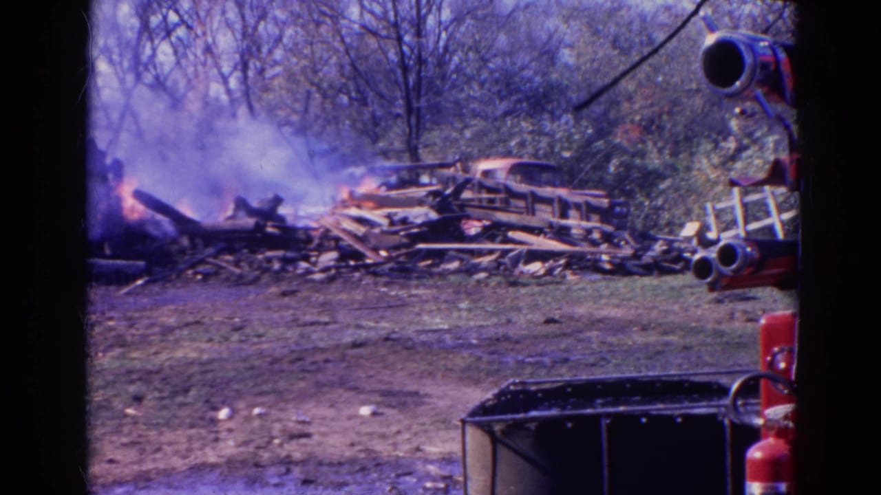 Burning debris pile with smoke, red fire equipment partly visible at the right edge.