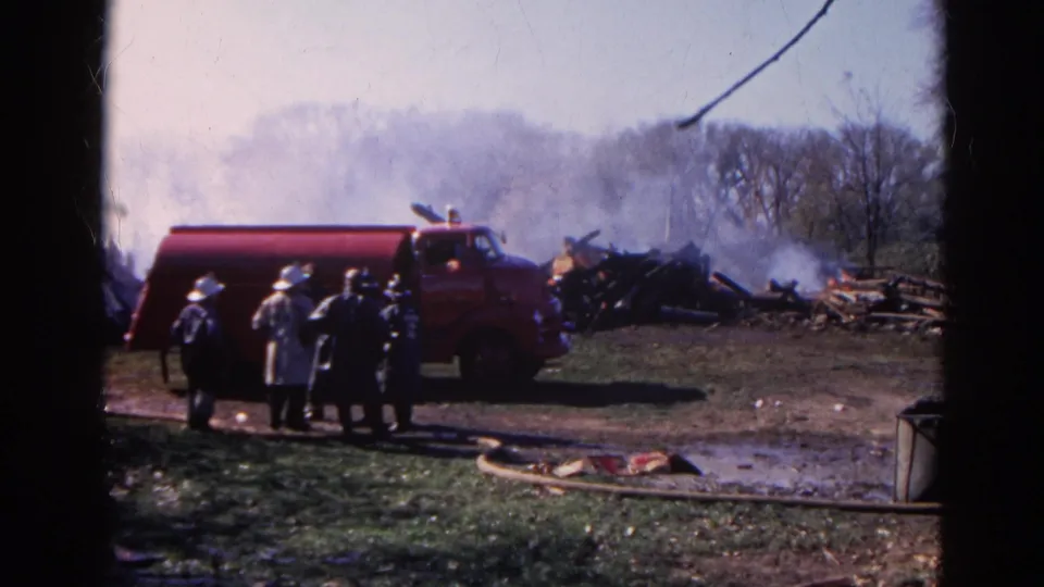 1968 Wauconda home-movie frame with firefighters in helmets gathered near a red fire truck and smoking wreckage.