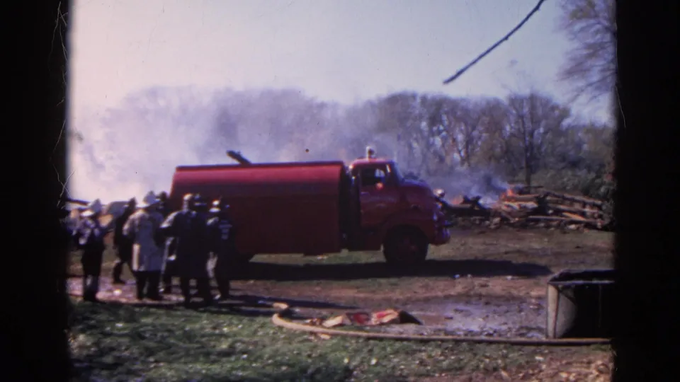 1968 Wauconda home-movie frame of a red fire truck parked near firefighters, hoses, and a smoky debris pile.