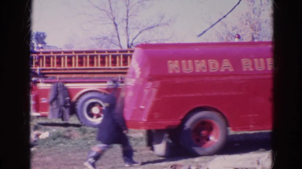 1968 Wauconda-area frame showing red rural fire trucks, a ladder truck, and a dark-coated firefighter walking through the field.
