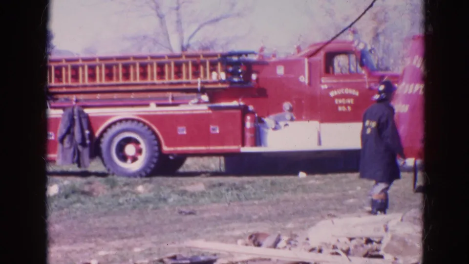 1968 Wauconda-area home-movie frame showing a red ladder truck and firefighters beside parked fire equipment in a grassy field.