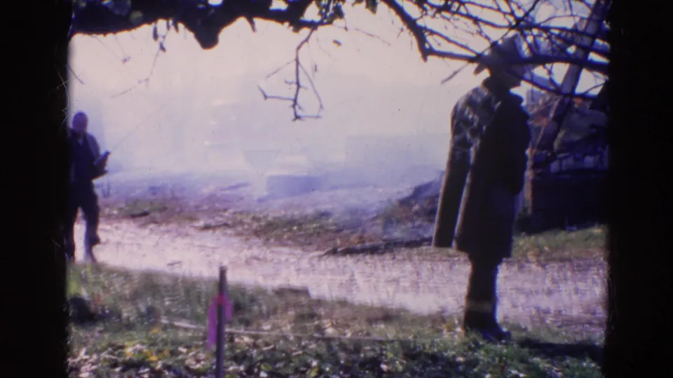 1968 Wauconda-area frame showing firefighters in dark coats watching smoke drift through a rural outdoor fire scene.
