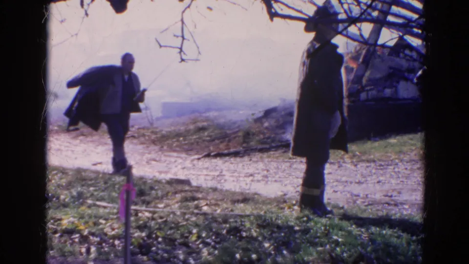 1968 Wauconda, Illinois frame with a man carrying a coat toward a second man in a long coat near smoke and scattered debris.