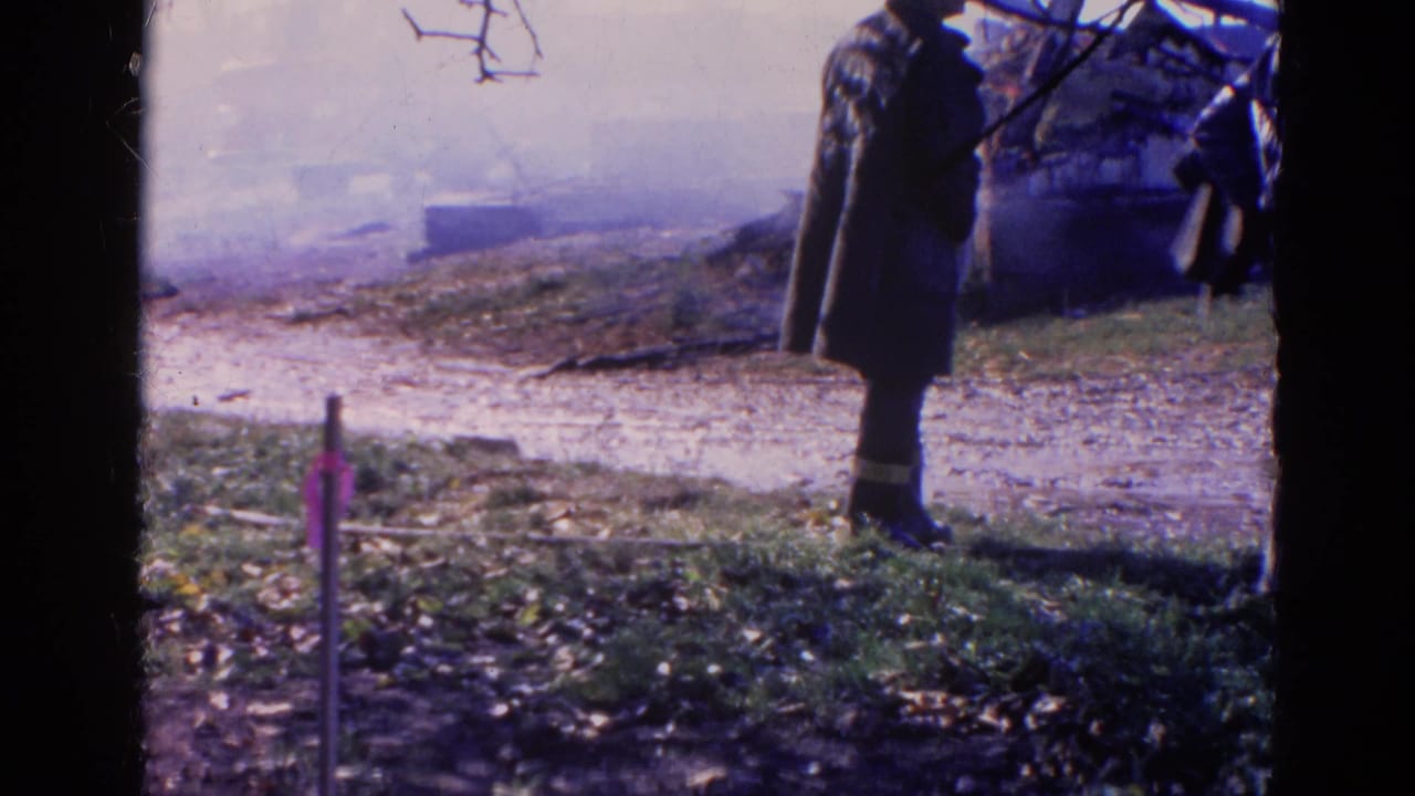 A person in a dark coat stands near smoke and burned debris behind a dark window frame.