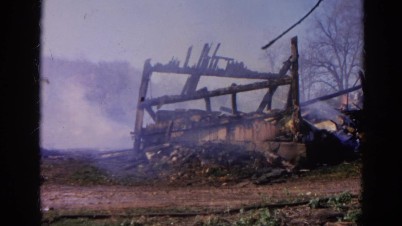 Smoke drifts around the charred frame of a burned wooden structure in an open lot.