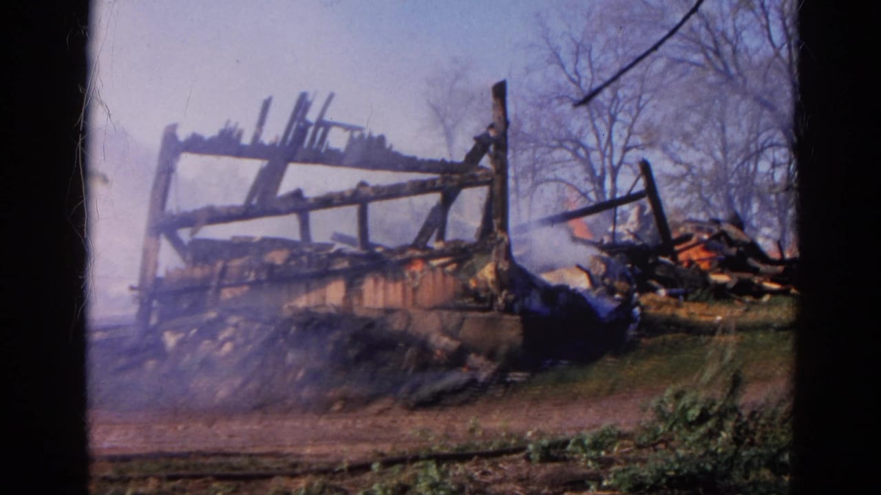 Charred wooden structure smolders with smoke and small flames behind an 8mm film border.