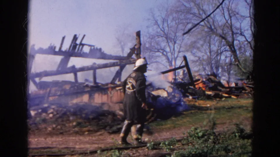 1968 Wauconda, Illinois 8mm frame showing a firefighter in dark gear beside smoke, debris, charred framing, and flame.