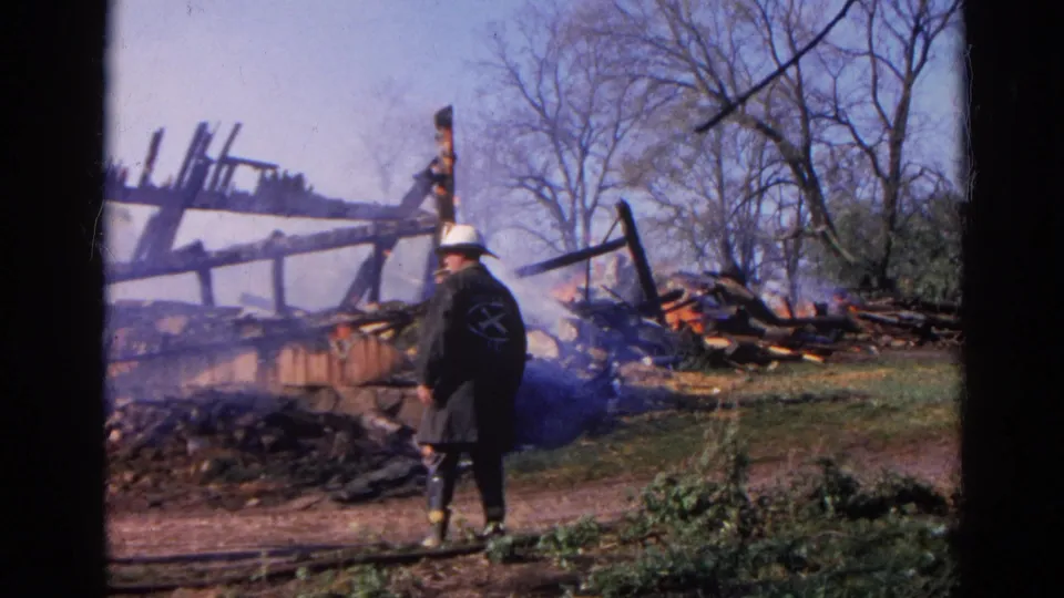 1968 Wauconda, Illinois frame showing a firefighter seen from behind near smoke, charred wood, rubble, and leafless trees.