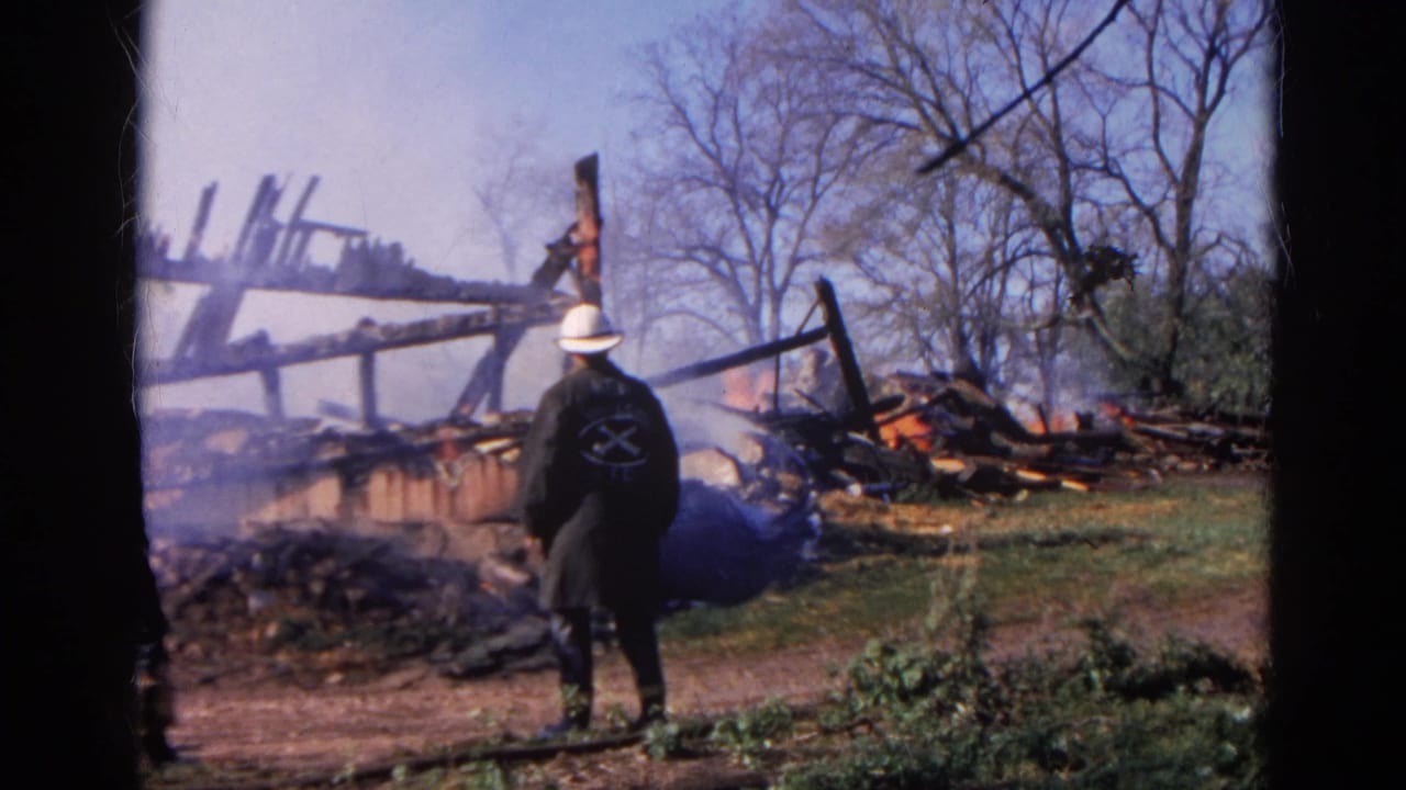 1968 Wauconda, Illinois 8mm frame showing a white-helmeted firefighter facing smoldering wreckage with flames and bare trees behind.