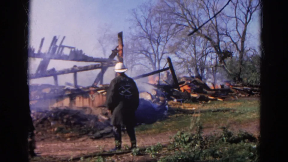 1968 Wauconda, Illinois 8mm frame showing a white-helmeted firefighter facing smoldering wreckage with flames and bare trees behind.