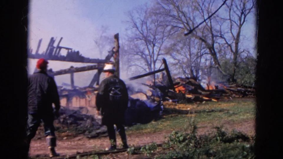 1968 Wauconda fire scene with a red-capped man and white-helmeted firefighter near collapsed smoking beams and visible flames.