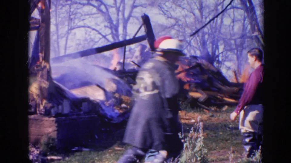 A white-helmeted firefighter crosses the foreground as a man watches smoking fire debris in 1968 Wauconda, Illinois footage.