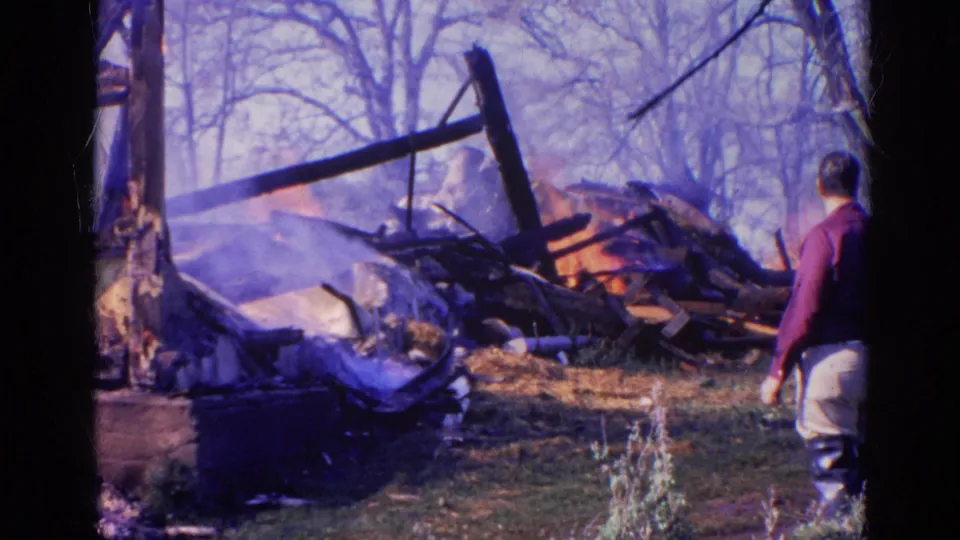 1968 Wauconda fire aftermath frame showing a man beside smoking wreckage, charred beams, scattered debris, and bare trees.