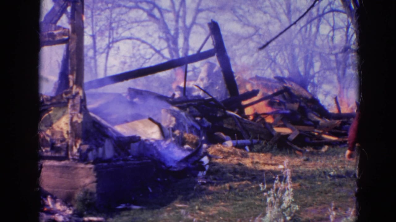 1968 Wauconda 8mm frame showing burned wooden debris, haze, bare trees, and part of a person near the wreckage.