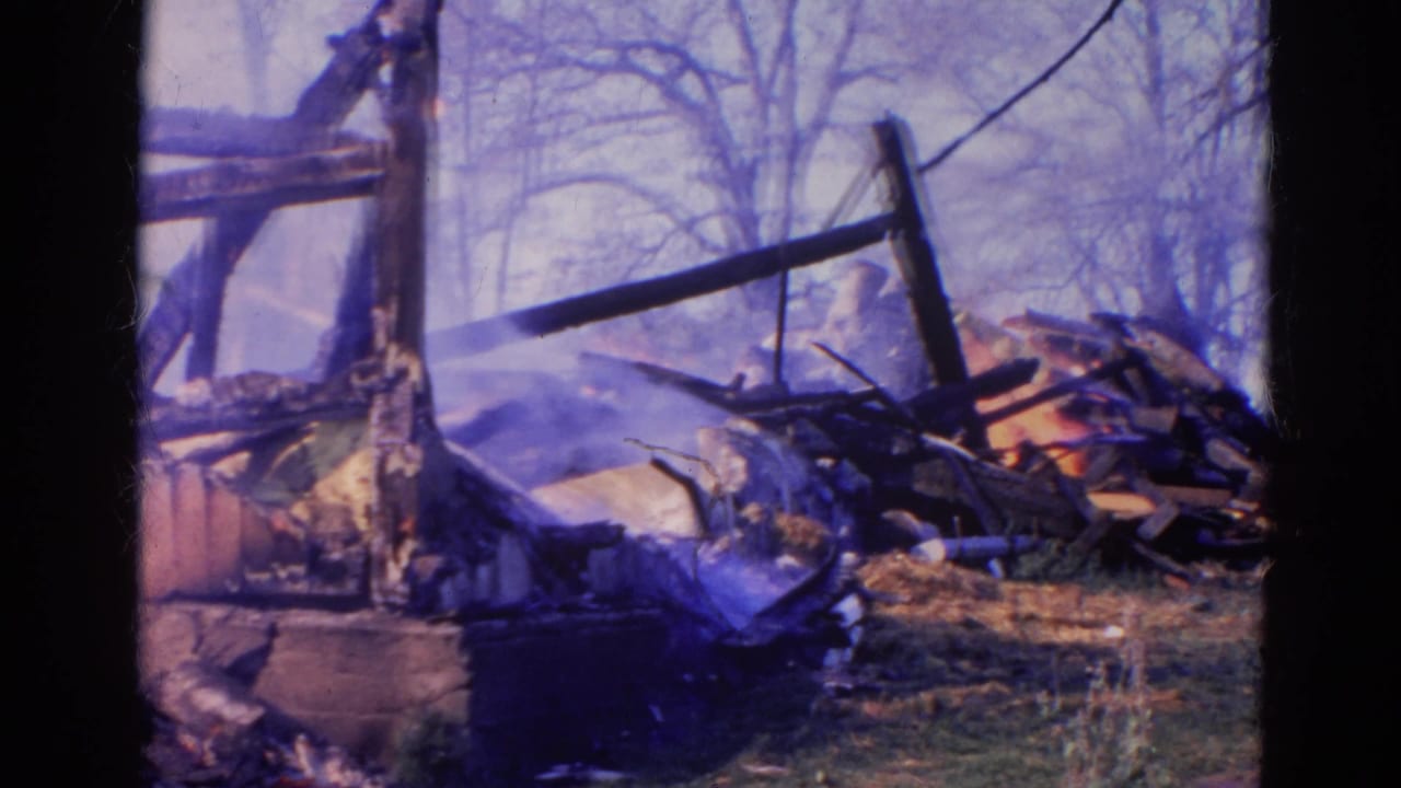 1968 Wauconda frame with collapsed fire debris, scorched wood, bare trees, and a partly visible person in red nearby.