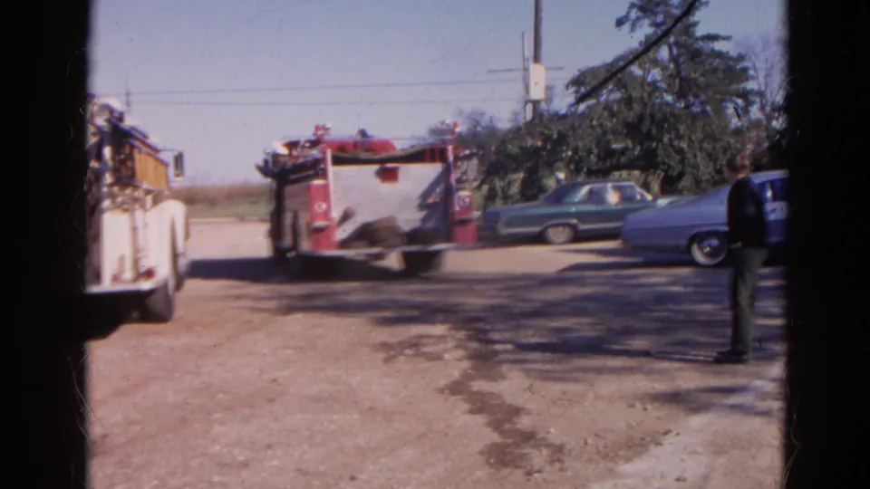 1968 Wauconda frame with a fire truck, several parked American cars, trees, utility wires, and a dirt roadside area.