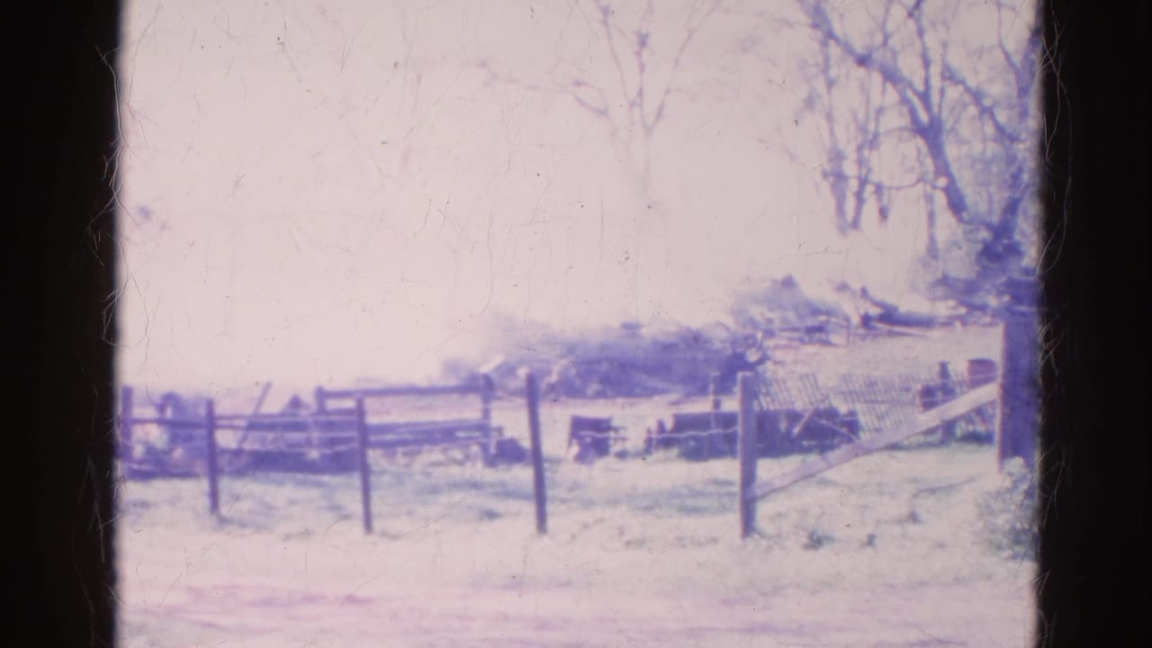 Smoke drifts behind a wooden fence near bare trees in a faded 8mm farm scene.