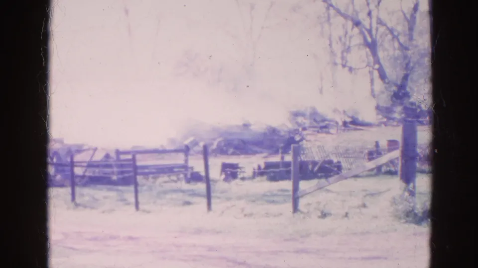 1968 8mm frame from Wauconda showing a smoky burn area behind a wooden fence and leafless trees.