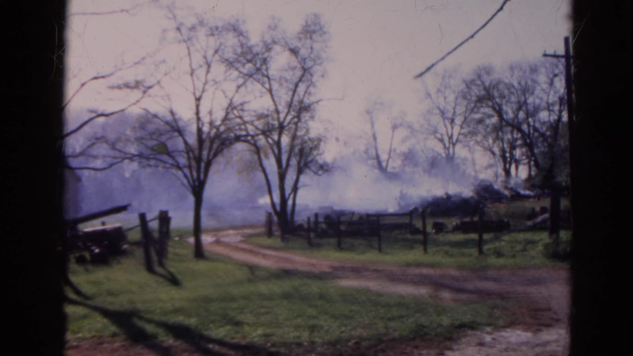 Rural dirt road beside grass and fencing, with smoke rising behind bare trees.