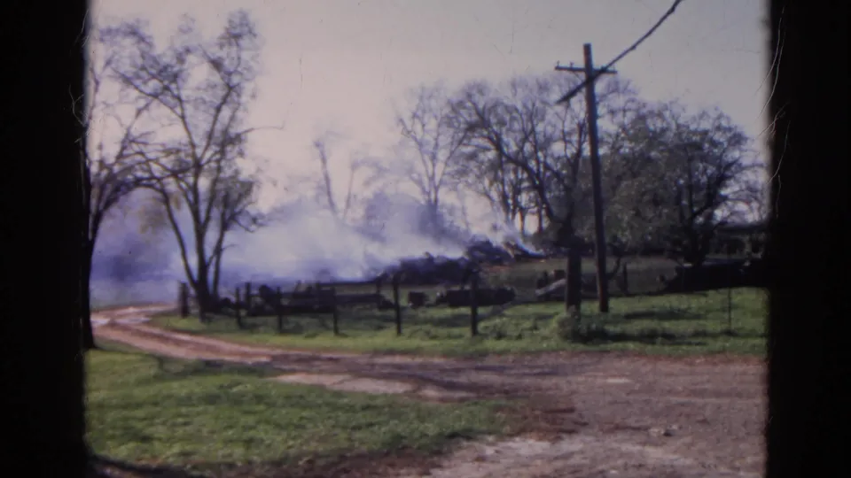 1968 Wauconda, Illinois home-movie frame showing drifting smoke beyond a fence, bare roadside trees, a utility pole, grass, and a dirt road.