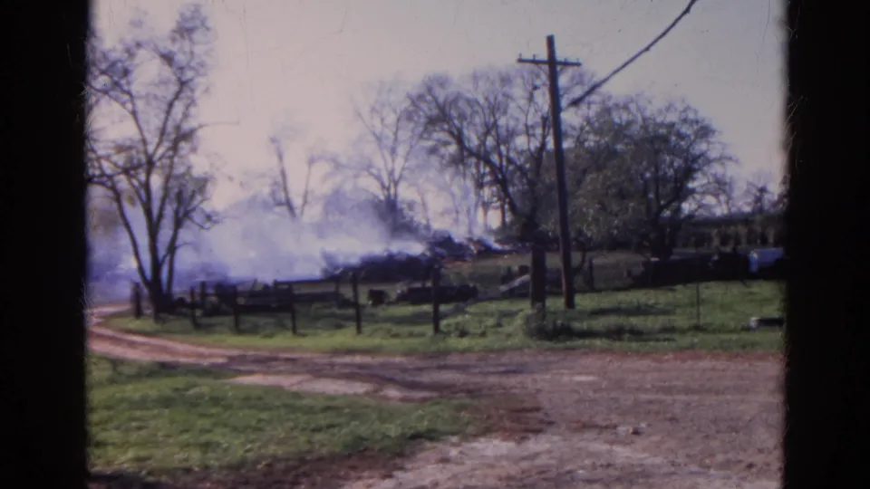 1968 Wauconda, Illinois frame with smoke beyond a fence, bare trees, utility poles, grassy roadside, dirt surface, and dark vehicle-like shapes at right.