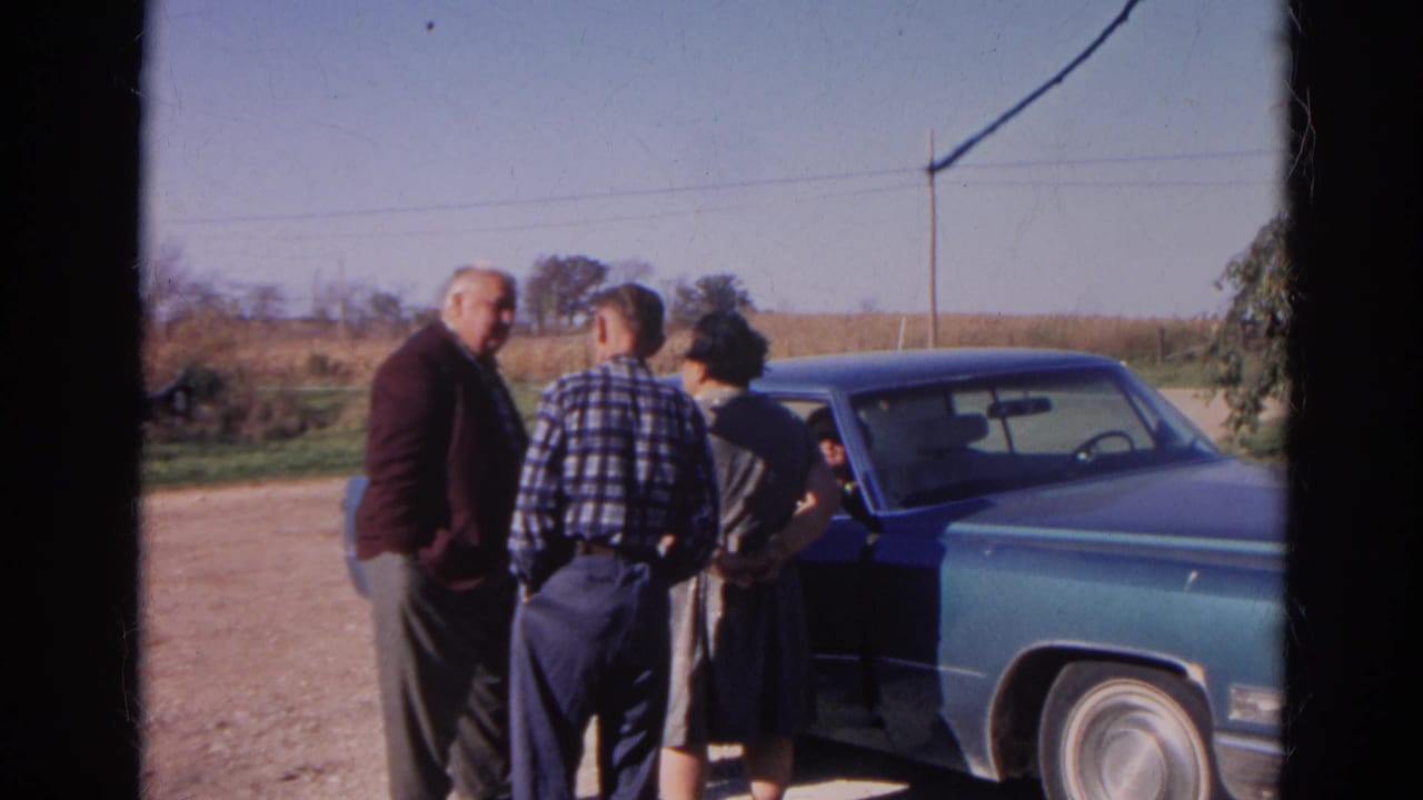 In the supplied 1968 Wauconda, Illinois home-movie frame, three adults stand near a blue sedan beside fields and a dirt road.