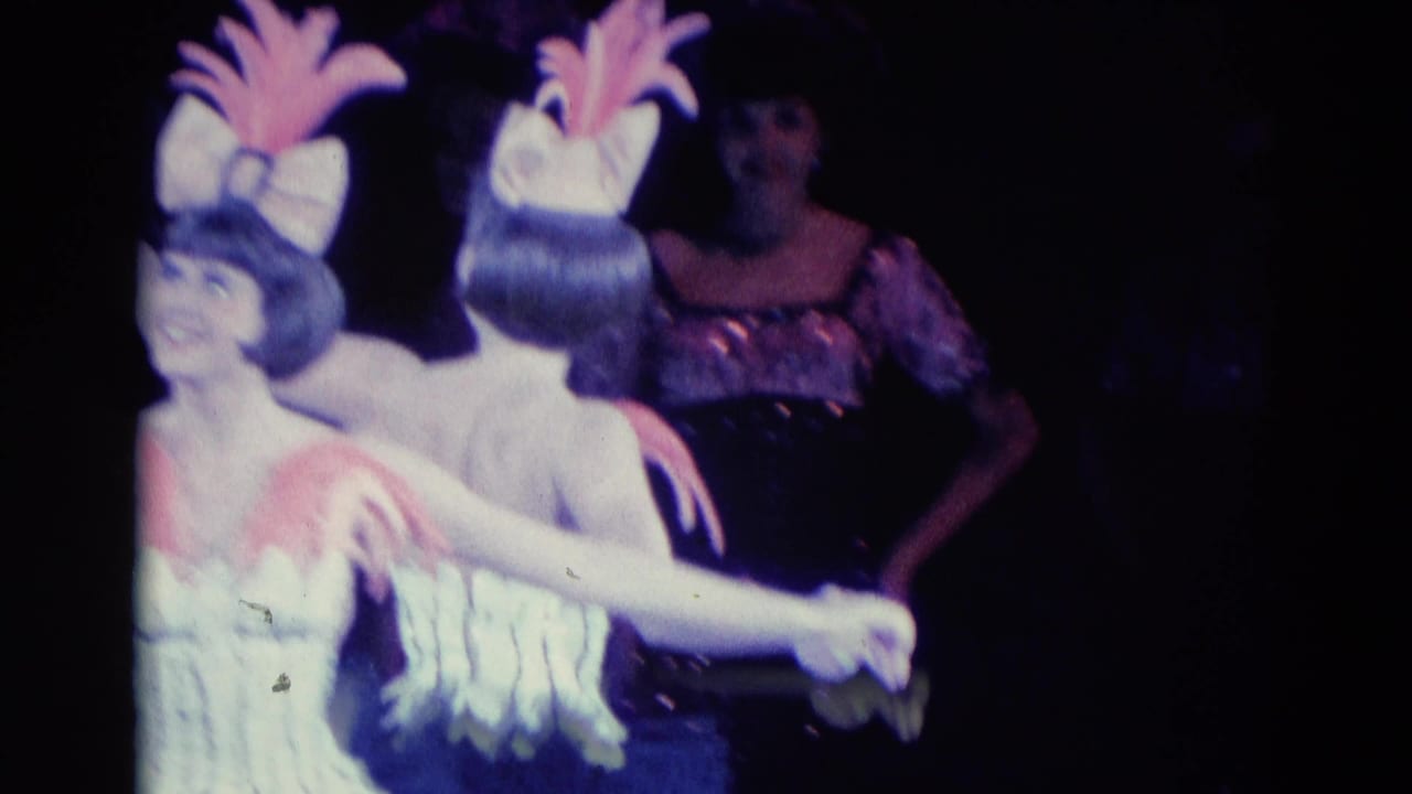 Two women in matching feathered stage costumes hold hands during a bow or turn on a dark stage.