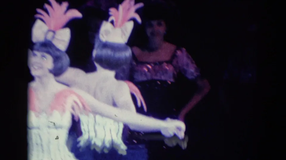 In 1969 Chicago home-movie footage, two women in matching white and pink feathered costumes hold hands under dim stage lighting.