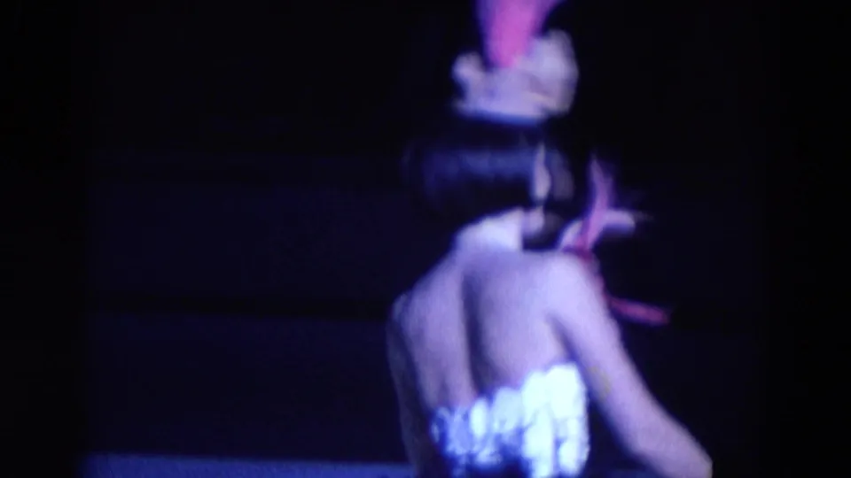 A costumed performer turns away from the camera on a dim stage, with the feathered headpiece and white outfit visible in 1969 Chicago footage.