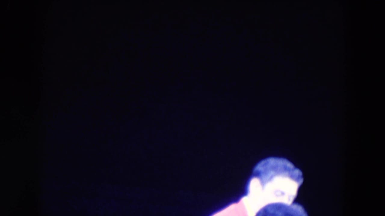 A male-presenting skater in a pink jacket is partly framed at the edge of a dark rink in 1969 Chicago footage.