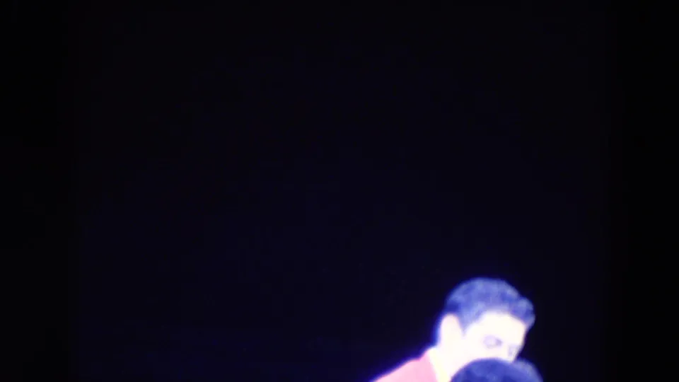 A male-presenting skater in a pink jacket is partly framed at the edge of a dark rink in 1969 Chicago footage.