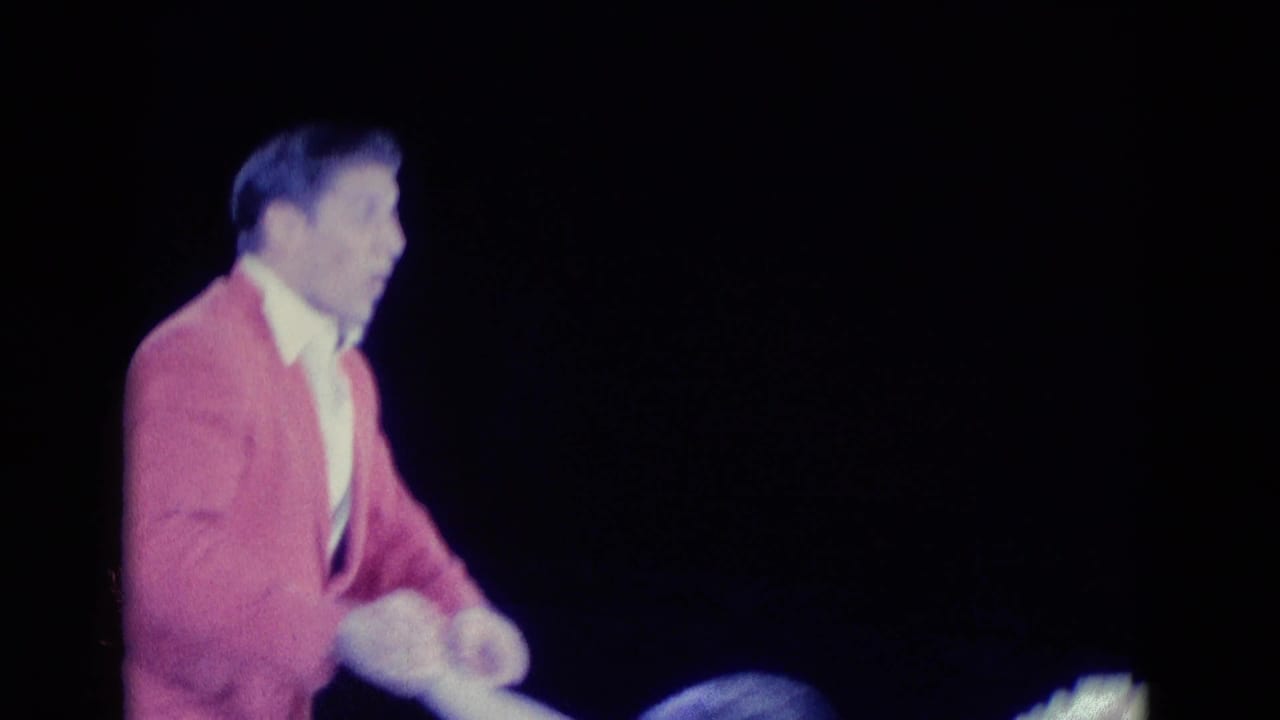 Male skater in a pink jacket moves across a dark ice rink.