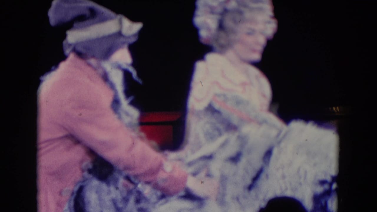 Costumed performers interact on a dark stage with red lights behind them.