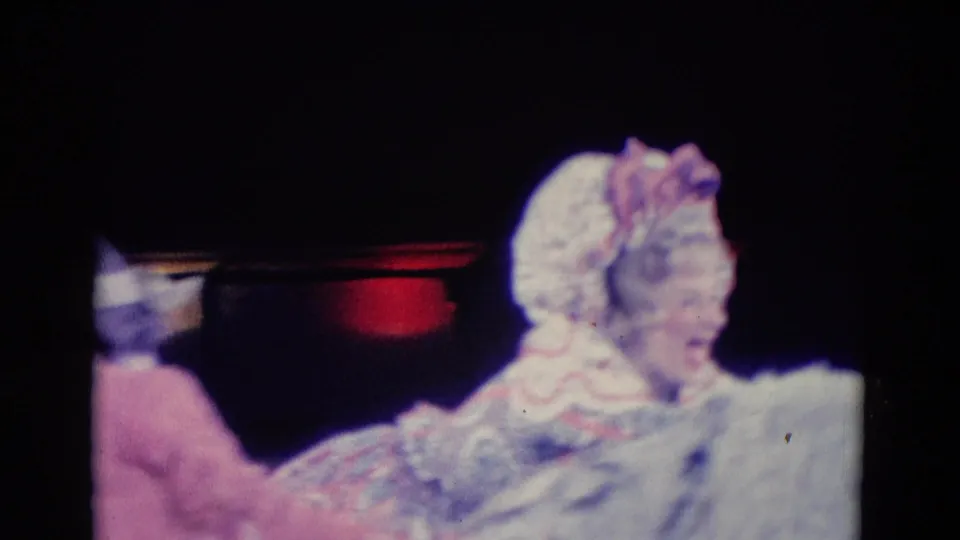 A pale-costumed performer is visible beside a white animal figure, with another performer partly seen at the left edge.