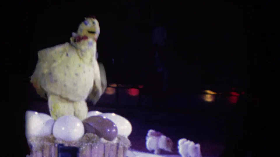 1969 Chicago 8mm frame of a spotted chicken puppet on a small stage nest with large white and purple eggs below.