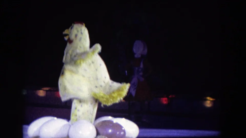 1969 Chicago stage frame showing the chicken puppet turned sideways near oversized eggs, with costumed performers partly visible in shadow.