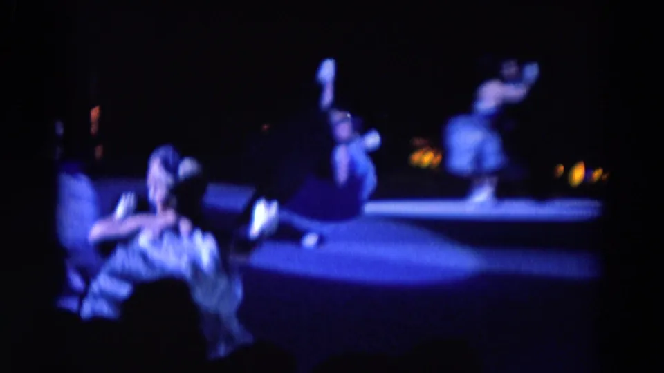 1969 Chicago home-movie frame showing several costumed skaters on a dim ice rink with blue theatrical light and blurred audience silhouettes.
