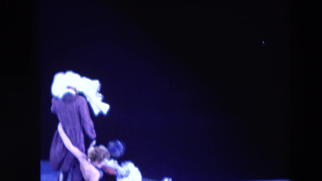 1969 Chicago ice-show frame with a dark-costumed performer wearing a large white headpiece beside another skater under blue light.