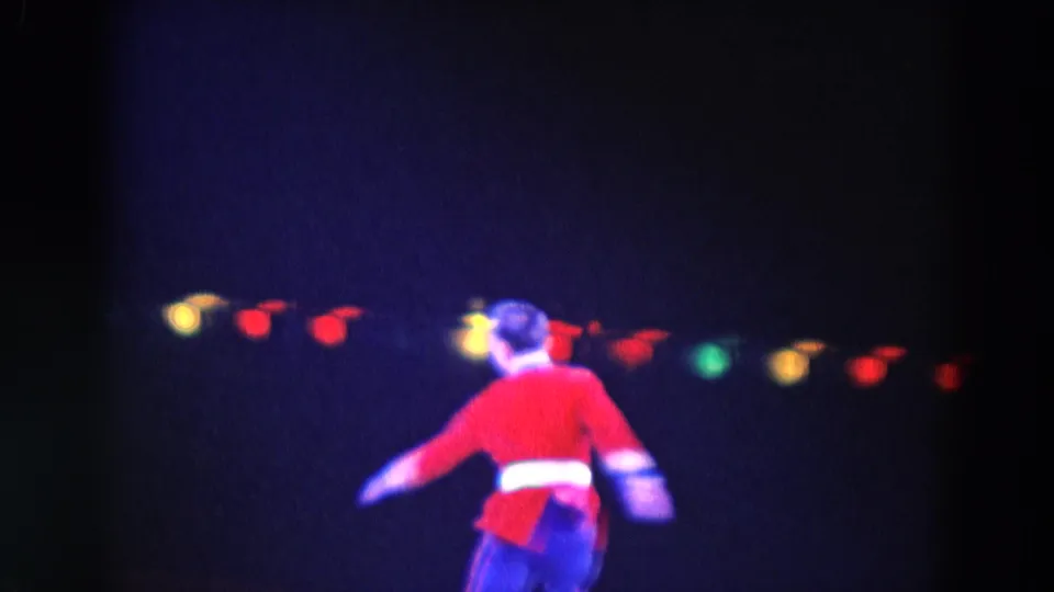 1969 Chicago home-movie frame showing a skater in a red jacket gliding across a dark rink with small colored lights behind him.