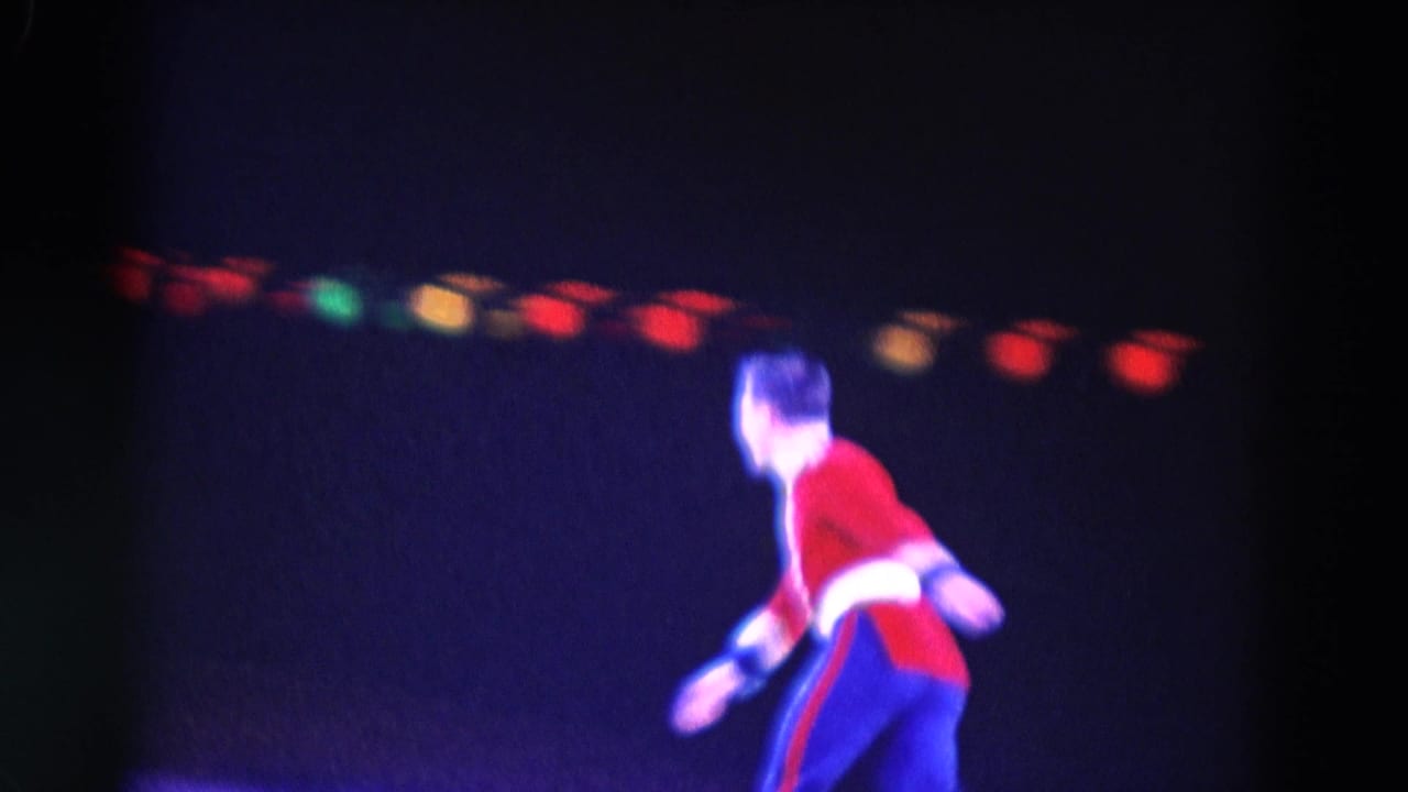 1969 Chicago home-movie frame showing a red-and-blue costumed figure skater moving across a dark ice rink under colored lights.