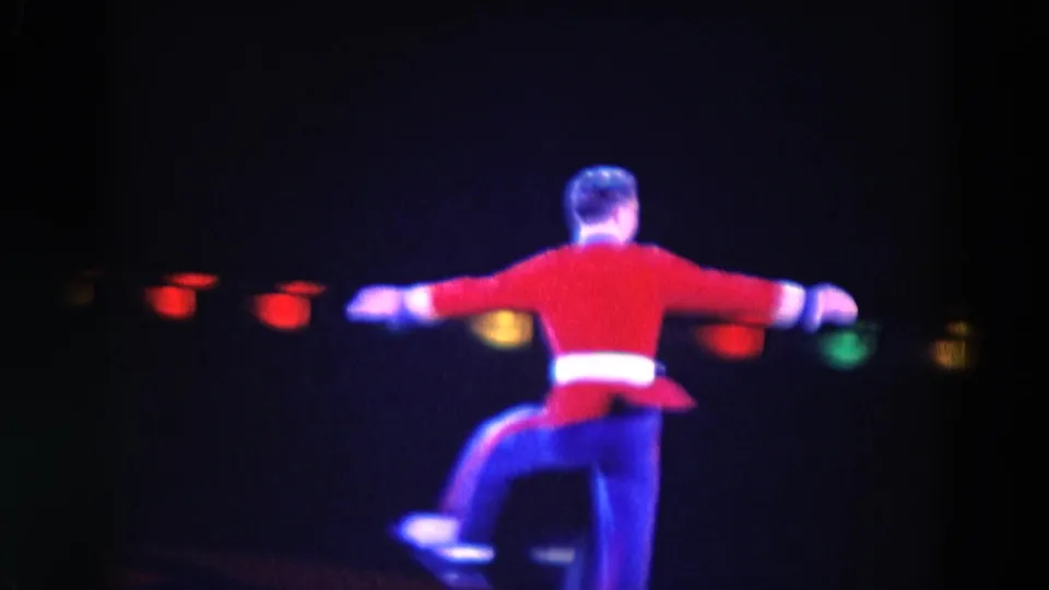 1969 Chicago home-movie frame showing the costumed skater from behind, arms extended during a turn or spin.