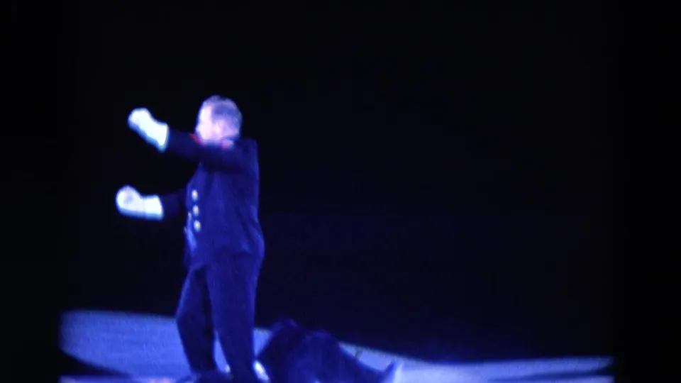 1969 Chicago skating-show frame showing a dark-clad performer or announcer gesturing with white gloves on the rink.