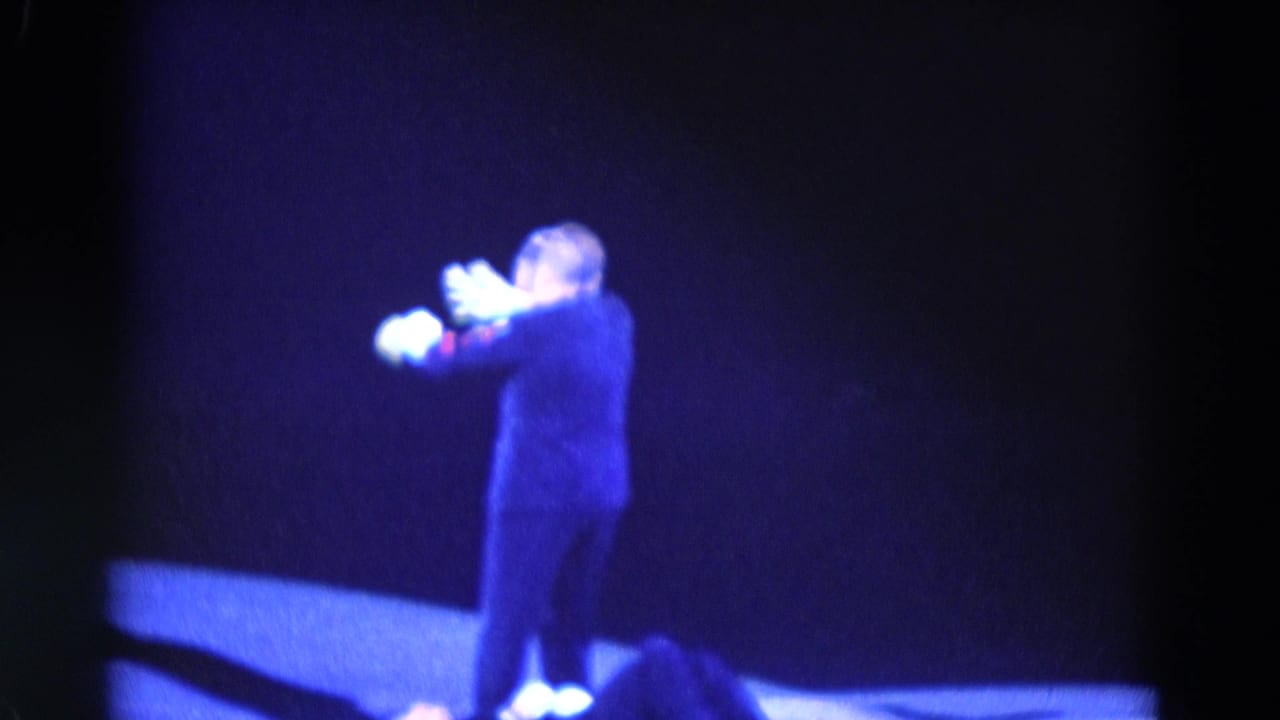 Dark-suited performer gestures with white gloves near a person lying on the ice.