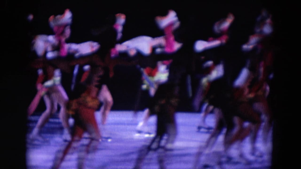 In 1969 Chicago home-movie footage, ensemble dancers appear in formation on a dark stage with saturated purple-blue light.