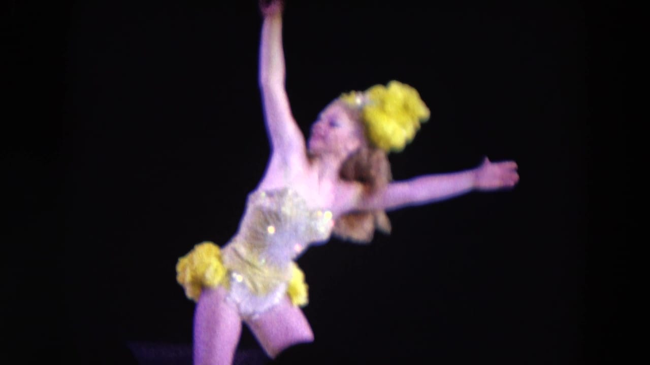 A solo stage dancer in a sparkling yellow and gold costume extends her arms against a dark background in 1969 Chicago footage.