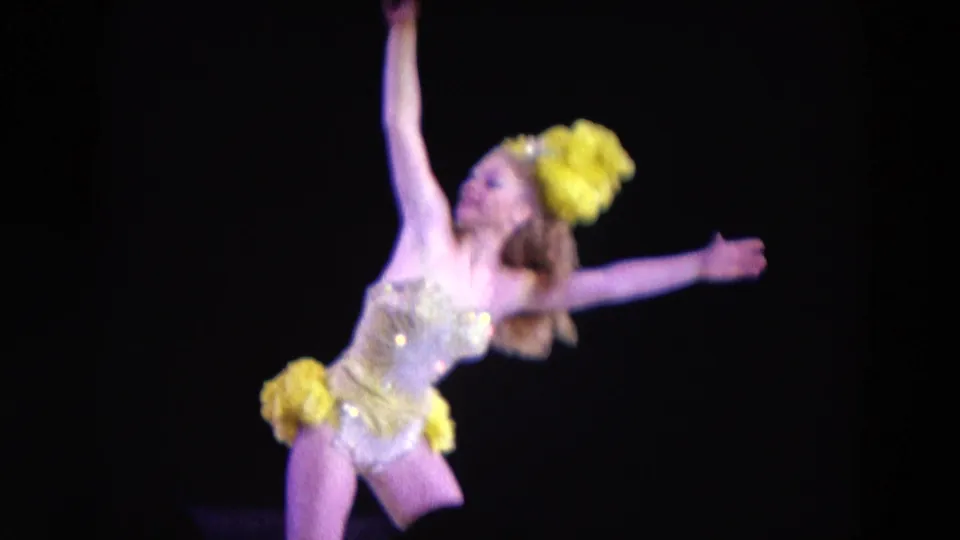 A solo stage dancer in a sparkling yellow and gold costume extends her arms against a dark background in 1969 Chicago footage.