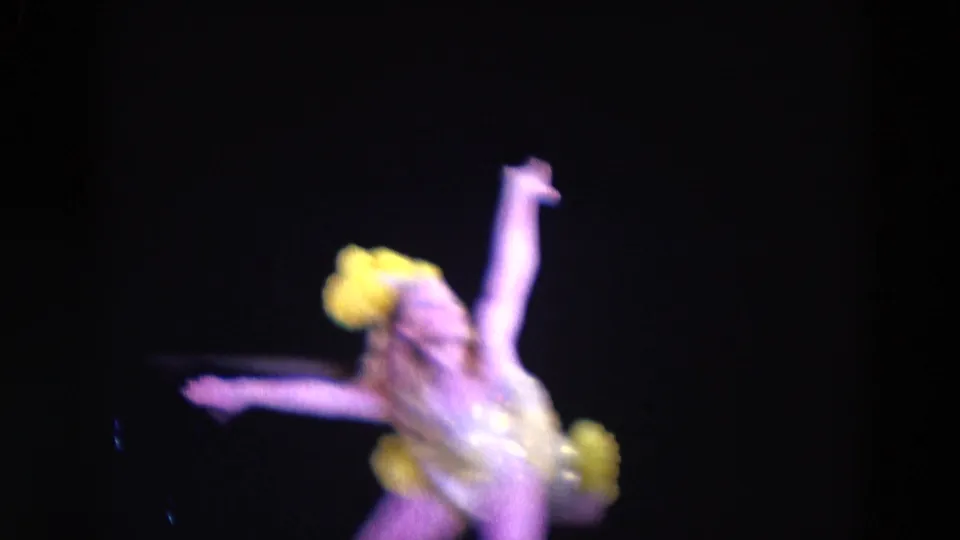 The solo performer in yellow and gold appears blurred in motion, with one arm raised and stage light reflecting off the costume.