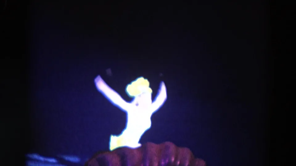 A strongly overexposed solo dancer in a yellow costume raises both arms, with a darker foreground form partly visible below.