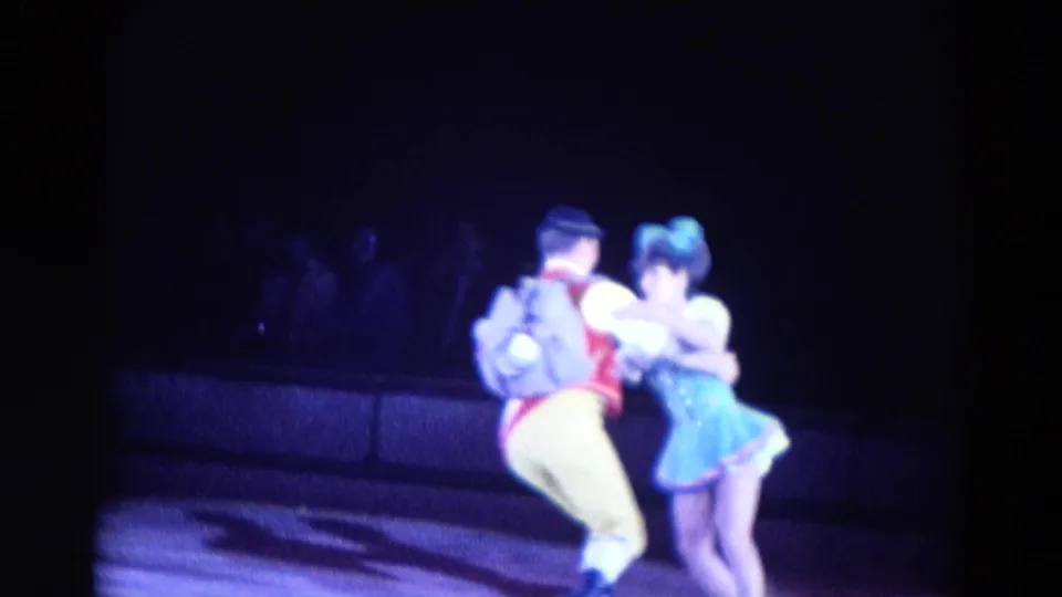 A male skater in a red vest and yellow pants partners a female skater in blue near the boards in 1969 Chicago rink footage.